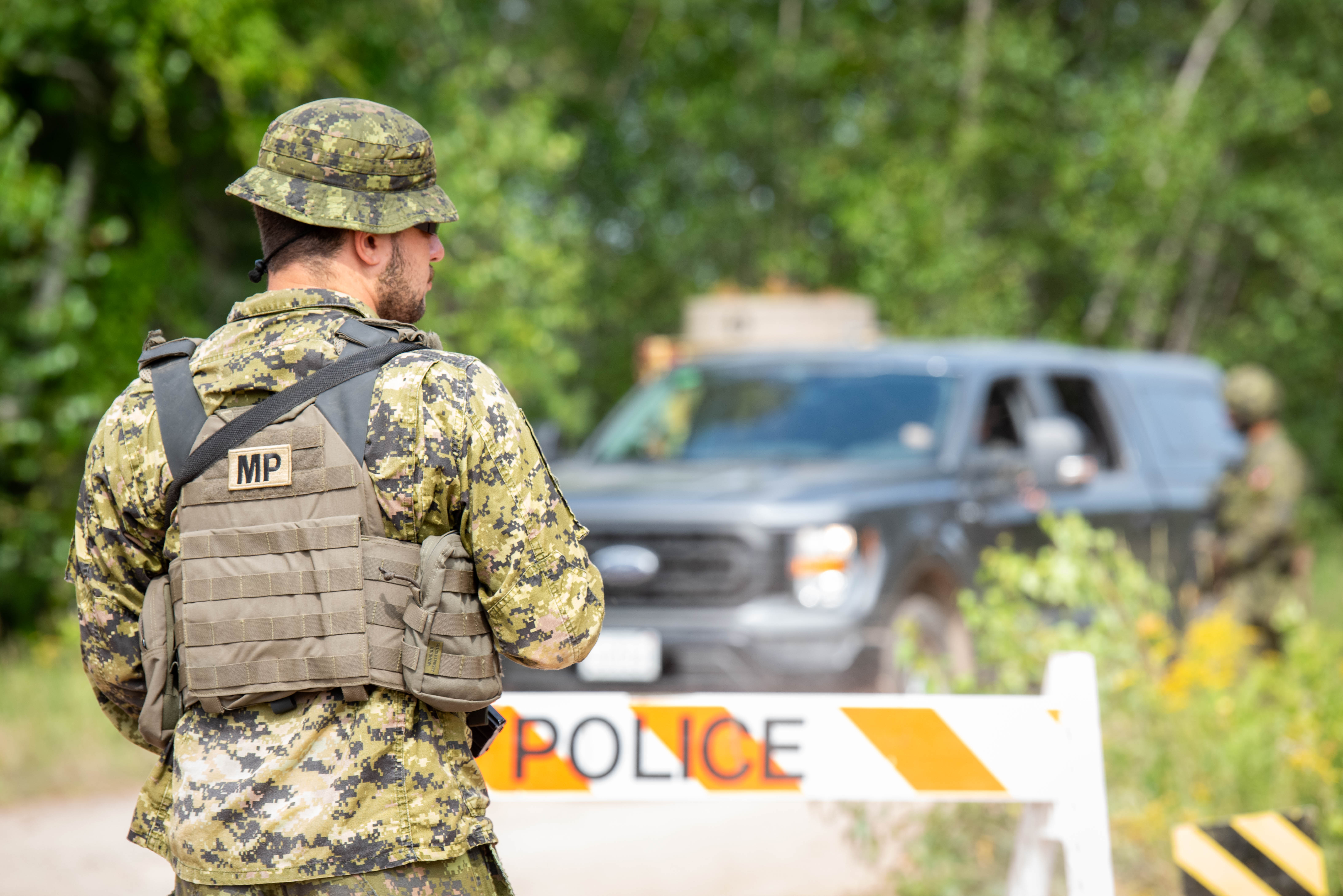 A Military Police member is stood in front of an orange and white striped barrier with “Police” 
written on it. In the background is a grey vehicle and trees. 